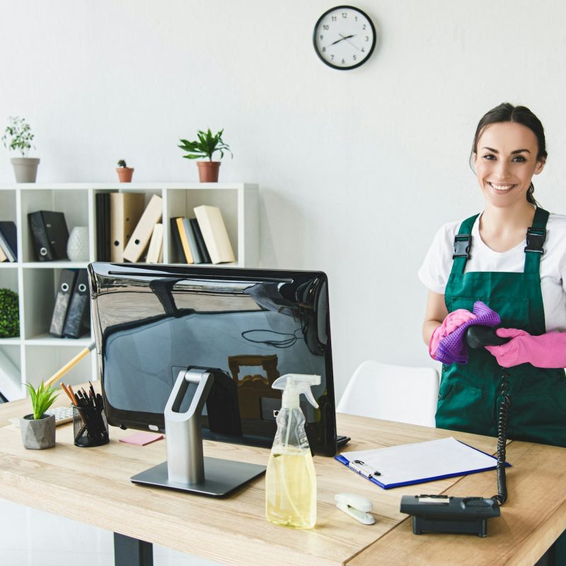 beautiful young professional cleaner smiling at camera while cleaning modern office